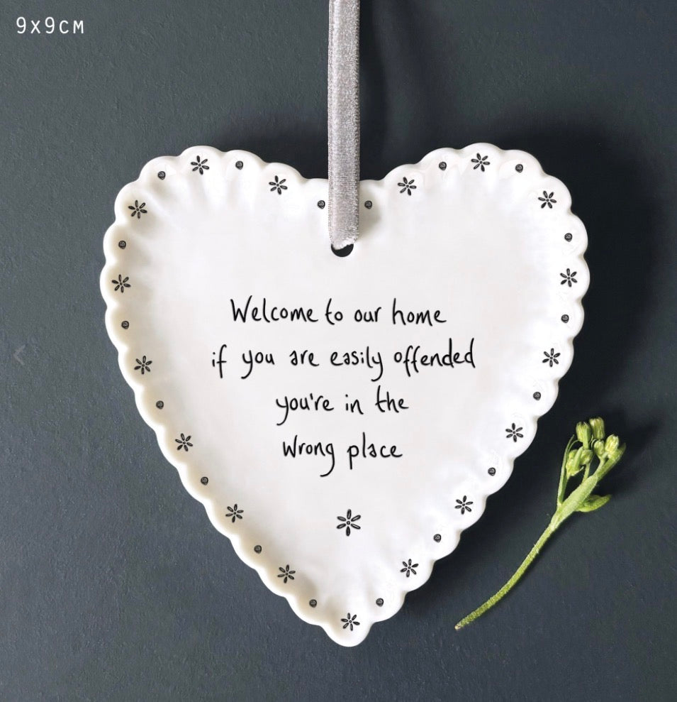 White East of India porcelain hanging heart with a scalloped daisy border, delicate black floral detail, and the humorous sentiment "Welcome to our home if you are easily offended you're in the wrong place", displayed with a grey satin ribbon.