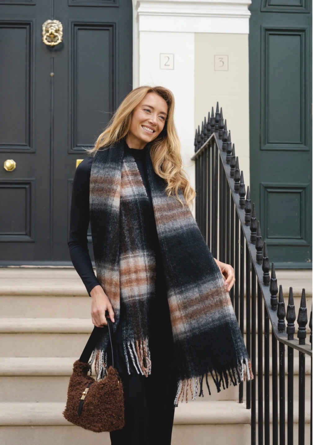 Oversized plaid check blanket scarf in charcoal black, brown, tan, and cream tones with long tassel fringe ends, draped as a cosy winter wrap.