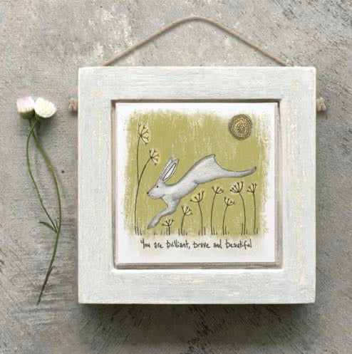 East of India small square whitewashed wooden framed picture of a leaping white hare on a sage green background with wildflower stems, a gold sun circle, and the sentiment "You are brilliant, brave and beautiful," hung with natural jute string.