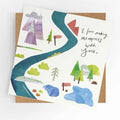 Square illustrated greeting card with a soft watercolour and paper collage landscape scene featuring a winding river path, mountains, trees, signpost, and red mailbox in teal, green, purple, red, and grey tones, with handwritten-style script reading "I love making memories with you," shown alongside a kraft brown envelope.