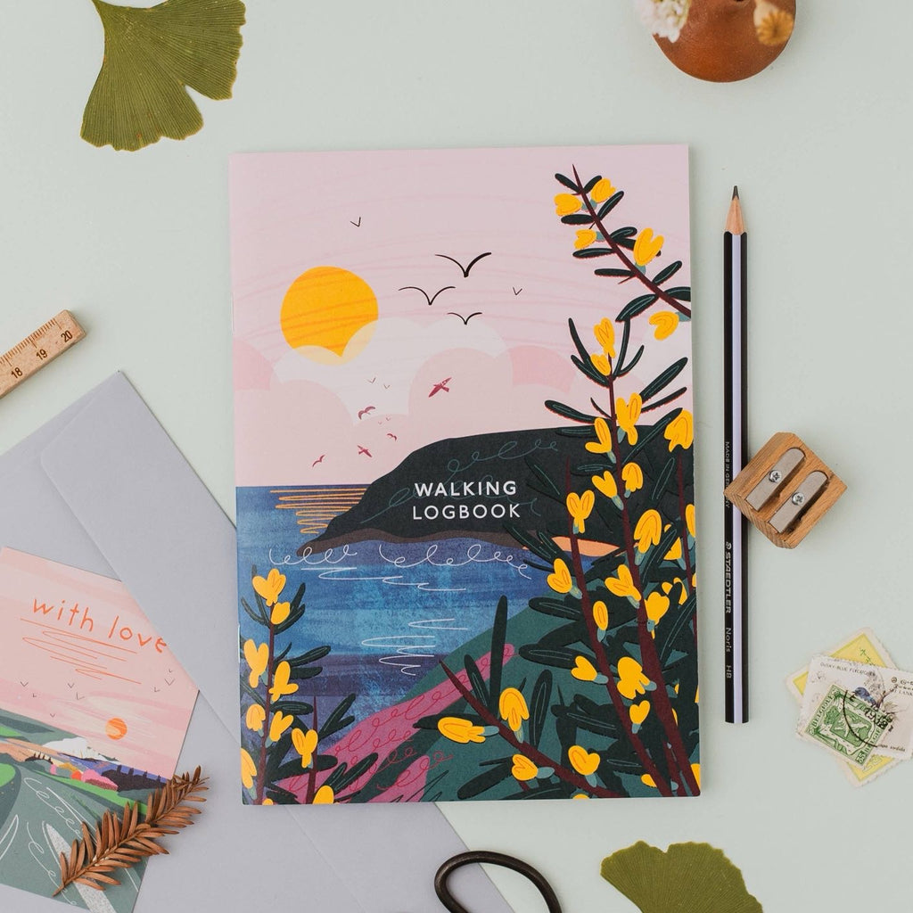 The Walking Logbook by Onneke featuring an illustrated cover with yellow gorse flowers, a dark coastal headland, blue sea, and a soft pink sunrise sky with birds in flight.