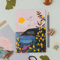 The Walking Logbook by Onneke featuring an illustrated cover with yellow gorse flowers, a dark coastal headland, blue sea, and a soft pink sunrise sky with birds in flight.