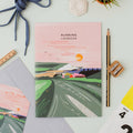 The Running Logbook by Onneke, featuring a beautifully illustrated cover with a winding running path through rolling green hills, a bridge, a coastal town, and a pink sky with a warm orange sun.