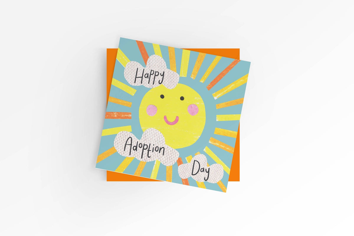Square Happy Adoption Day greeting card featuring a smiling illustrated sun with yellow and orange rays and three fluffy white clouds on a blue sky background, shown with a bright orange envelope.