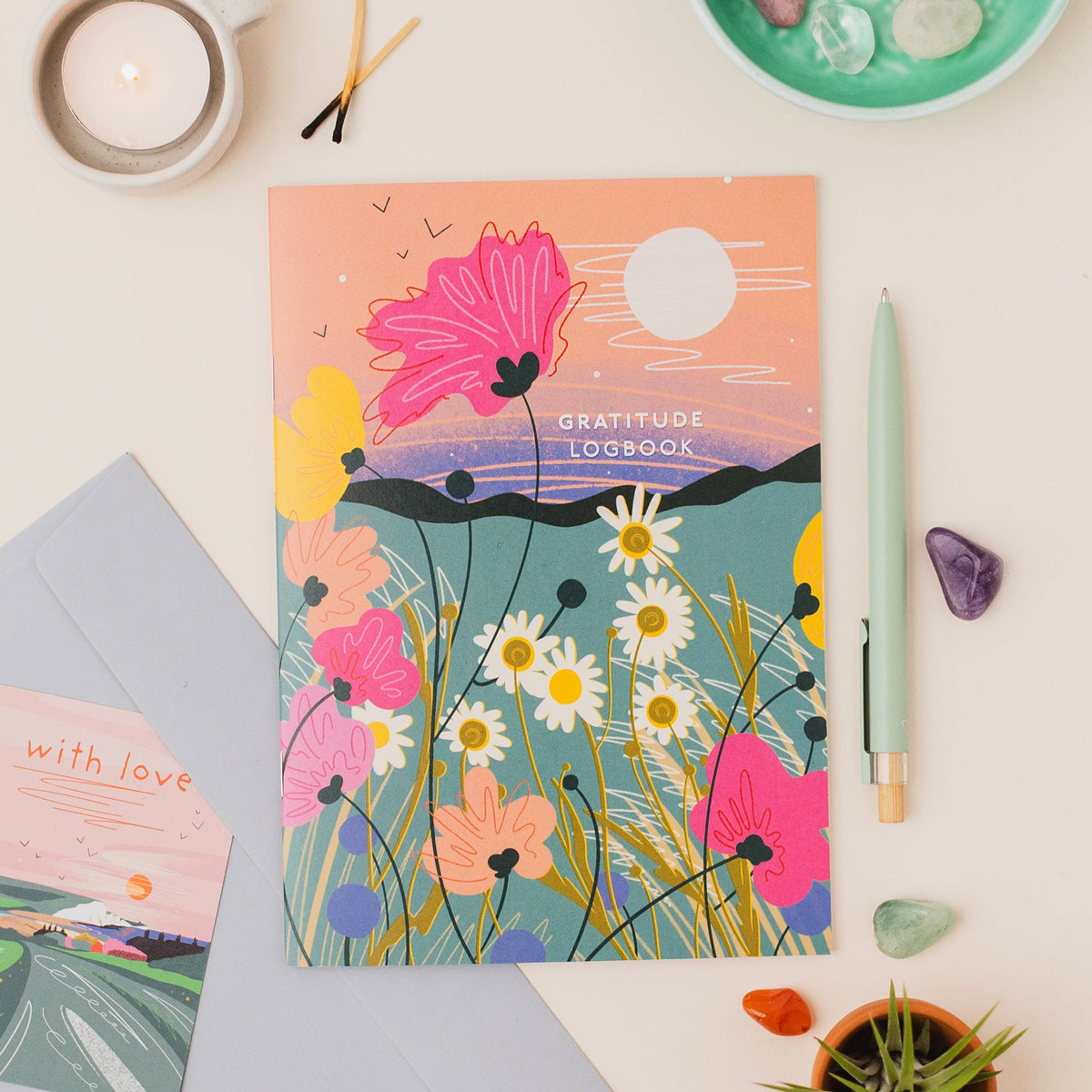 Onneke Gratitude Logbook in vibrant wildflower colourway with hot pink poppies, white daisies, peach and lavender blooms, and golden grass stems against a warm peach and orange sunset sky with a full white moon.
